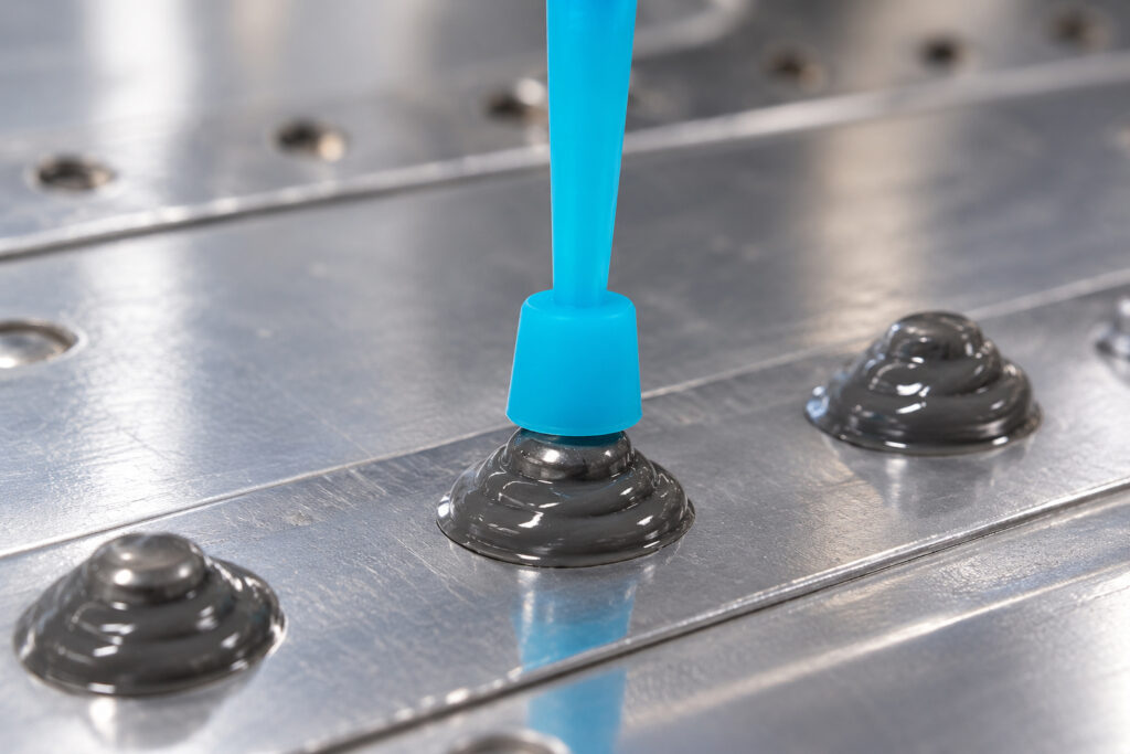 Close-up of a blue sealant application nozzle dispensing gray aerospace sealant onto an aircraft rivet on a metallic panel, ensuring precise sealing of aircraft fasteners.