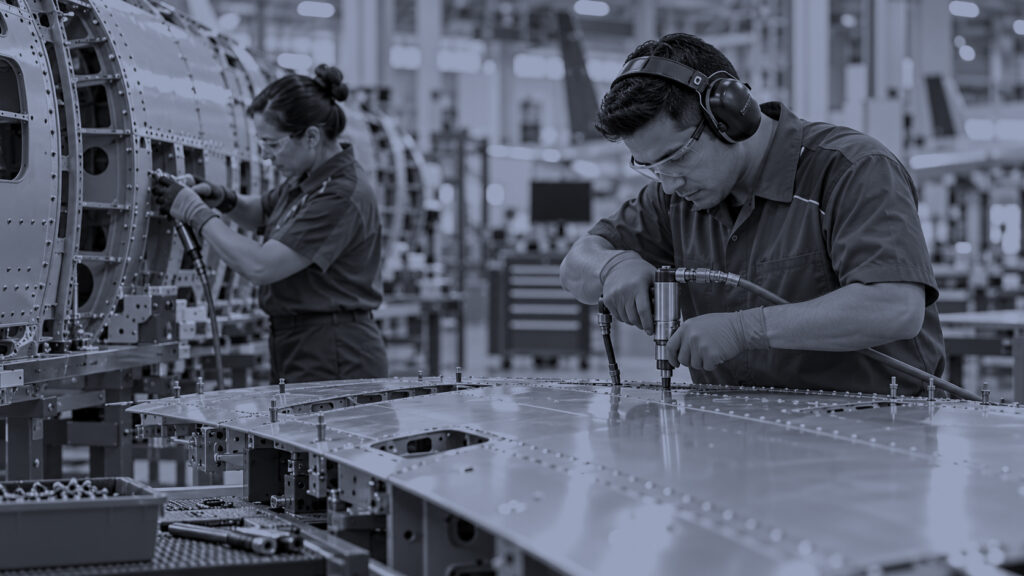 Two skilled aerospace technicians working on aircraft structural components in a manufacturing facility, using precision tools on metal parts.